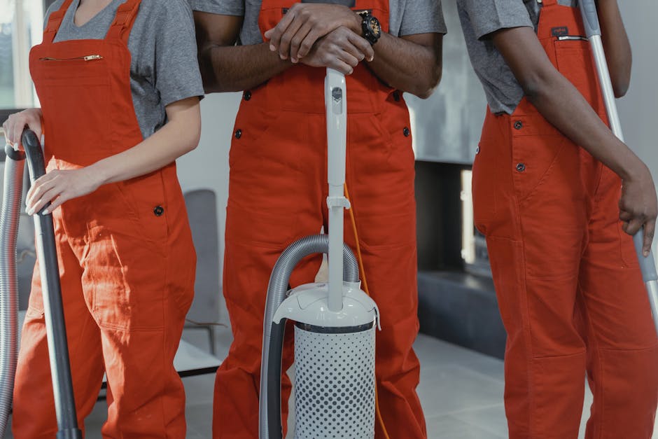 Close-up of three professional cleaners from Cleaners N1, dressed in grey t-shirts and bright orange overalls, performing surface cleaning with a vacuum cleaner in an indoor setting. The vacuum features a white and grey cylindrical body with a flexible grey hose, and it is actively used on the floor, which appears to be made of ceramic tiles. The room is well-lit, and the focus is on the tools and the cleaning process, emphasizing hygiene and maintenance of surfaces such as floors using modern cleaning equipment. The background includes blurred elements of a clean, modern interior space, highlighting the company's focus on deep cleaning and sanitisation services on Upper Street Islington, N1.