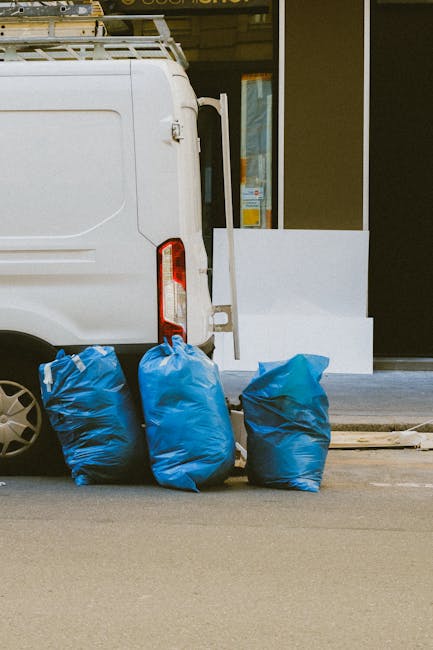 A close-up of the rear side of a white commercial van parked on a street in front of a modern building with glass doors and windows. The van features a vertical taillight, and the back door is partially visible. Several large blue garbage bags are positioned on the pavement beside the van, ready for waste disposal. The scene suggests waste collection or outdoor cleaning activity, with clean surfaces and a tidy arrangement. The setting is well-lit with natural daylight. This image reflects the cleaning and waste management services provided by Cleaners N1, as showcased on the page about cleaning services on Upper Street Islington, N1.