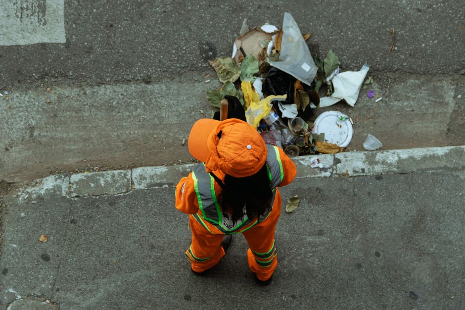 A street-level scene showing a sanitation worker from Cleaners N1, dressed in a bright green uniform and matching cap, standing next to a large green waste bin on a paved city sidewalk. The worker is